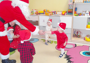 Święty Mikołaj wita się z dziewczynką w mikołajkowej czapeczce.