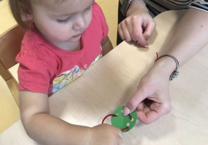 Opiekunka przytrzymuje Laurze zielony guzik podczas nauki nawlekania sznurka na guziki.