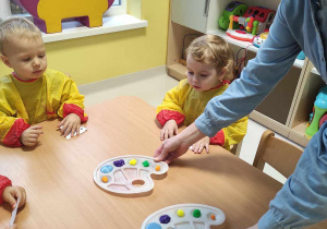 Opiekunka kładzie na stoliczkach dzieci dwie paletki z kolorowymi farbami.