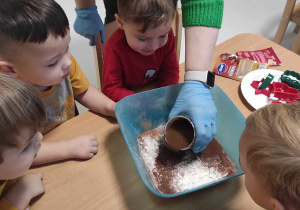 Dzieci przyglądają się jak opiekunka wlewa do miski z sypkimi składnikami roztopione masło z miodem.