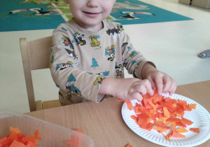 Uśmiechnięty Leon pozuje do zdjęcia podczas przyklejania na swój papierowy talerzyk kawałeczki pomarańczowej bibuły.
