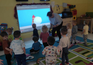 Dzieci i opiekunka naśladują wyświetlanego na tablicy interaktywnej chłopca, który toczy wielką kulę śnieżną.