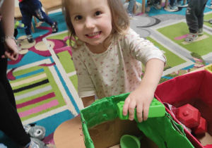 Uśmiechnięta Lilianna wkłada wybranego przez siebie zielonego klocka do tekturowego pudełka w tym samym kolorze.