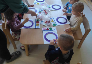 Zdjęcie opiekunki rozdającej dzieciom na stoliczki plastikowe pudełeczka z wyciętymi szablonami kolorowych ubrań.