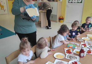 Opiekunka trzymając w dłoni papierowy talerzyk z ciastem do pizzy, tłumaczy dzieciom na czym będą polegać przeprowadzane zaraz zajęcia.