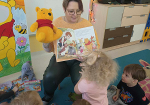 Zdjęcie opiekunki pokazującej dzieciom trzymaną w dłoni maskotkę oraz książeczkę Kubusia Puchatka.