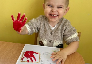 Uśmiechnięty chłopiec macha do zdjęcia rączką pomalowaną na czerwono.