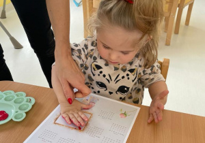 Opiekunka pomaga dziewczynce odcisnąć swoją rączkę na kalendarzu.