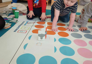 Miłosz i Szymon przyglądają się robotowi, który podczas kodowania rysuje pomarańczowym mazakiem kształt kwadratu.