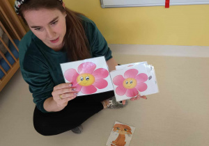 Opiekunka pokazuje dzieciom zalaminowane obrazki przedstawiające różowe uśmiechnięte kwiatuszki.