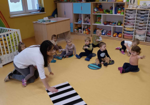 Zdjęcie opiekunki z grupy Motylki, tłumaczącej dzieciom przebieg przygotowanych na podłodze zajęć pt. ,,ZEBROWY MINI TOR''.