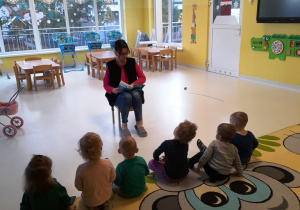 Opiekunka czytająca wiersz dzieciom siedzącym na dywanie.