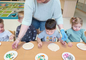 Opiekunka rozdaje dzieciom papierowe talerzyki z kropeczkami wykonanymi z kolorowej plasteliny.