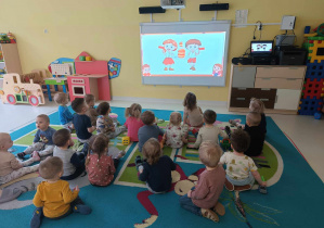Zdjęcie dzieci z grupy Pszczółki siedzących na dywanie, oglądających filmik edukacyjny o Tłustym Czwartku.