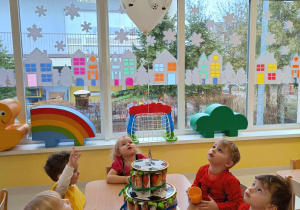 Dzieci spoglądają na wiszący biały balon przyczepiony do tortu wykonanego z owocowych musików.