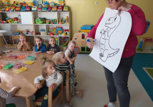 Opiekunka pokazuje dzieciom szablon dinozaura, który będą miały za zadanie ozdobić.