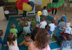 Zdjęcie dzieci z grupy Pszczółki siedzących na dywanie i przyglądających się opiekunkom trzymającym w dłoniach kolorowe tunele, które wykorzystane będą do zabawy pt. ,,DO JASKINI I NA SKAŁĘ''.