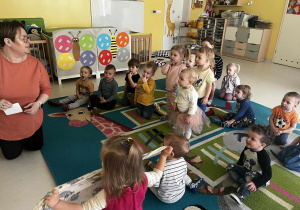 Zdjęcie dzieci siedzących i słuchających na dywanie opiekunki, która tłumaczy na czym polegać będą zaplanowane zajęcia.
