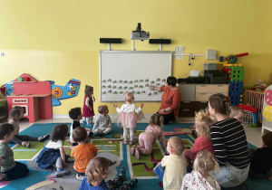Zdjęcie dzieci i opiekunek z grupy Pszczółki siedzących na dywanie przed tablicą interaktywną, przygotowujących się do zajęć pt. ,,ODSZUKAJ DINOZAURA''.