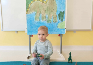 Dawid siedzący pod ścianką przedstawiającą zielonego dinozaura pozuje trzymając w dłoniach zielonego zabawkowego dinozaura.
