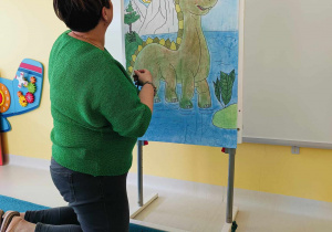 Opiekunka ustawia zabawkowe dinozaury na stelażu z narysowanym zielonym dinozaurem.