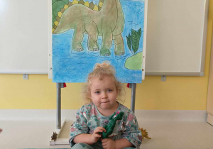 Laura siedząc ze skrzyżowanymi nogami pozuje do zdjęcia trzymając w rękach zabawkowe dinozaury.