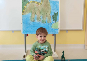Zdjęcie Tymoteusza siedzącego z zabawkowym brązowym dinozaurem pod ścianką fotograficzną.