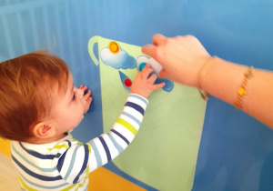 Opiekunka pomaga chłopcu przykleić chmurkę do garnka z papieru.