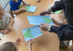 Opiekunka rozdaje dzieciom niebieskie kartki z przyklejonymi zielonymi listkami.