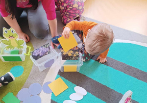 Zdjęcie Stanisława wkładającego do plastikowego pudełka z obrazkiem żonkili żółtą karteczkę w kształcie kwadratu.