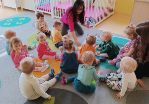 Zdjęcie dzieci i opiekunki z grupy Motylki siedzących na dywanie i przygotowujących się do zajęć pt. ,,WIOSENNE KOLORY''.