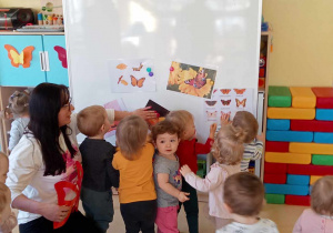 Zdjęcie dzieci i opiekunki z grupy Motylki stojących przy tablicy suchościeralnej i przygotowujących się do zajęć z okazji Dnia Motyla.