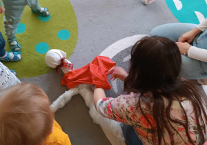 Dzieci przyglądają się jak jedna z opiekunek ubiera Marzannę w czerwoną sukienkę.