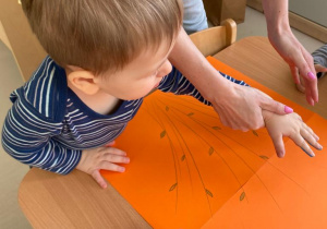 Opiekunka pomaga chłopcu odcisnąć swoją dłoń na pomarańczowym brystolu.
