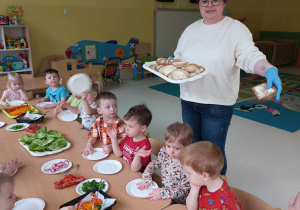 Opiekunka pokazuje dzieciom pokrojone i posmarowane masełkiem kromeczki chlebka.