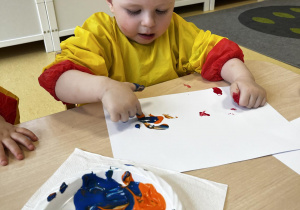 Mikołaj maluje na kartce paluszkiem namoczonym w pomarańczowo - niebieskiej farbie.