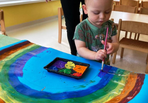 Mały chłopczyk maluje za pomocą pędzla tęczę na fioletowo.