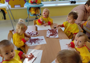 Dzieci z grupy Pszczółki malują farbami przy stoliku pnie drzew z rolki papieru.