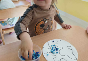 Zdjęcie zadowolonego Szymona podczas brania z plastikowego okrągłego pojemniczka bibułowych kulek w niebieskim kolorze.