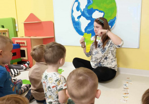 Opiekunka opowiada dzieciom o Planecie Ziemia, trzymając w dłoni szablon kontynentu i obrazek drzewka.