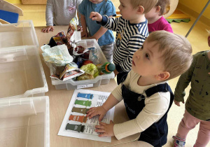 Zdjęcie dzieci z grupy Motylki oglądających rekwizyty znajdujące się w plastikowych prostokątnych pojemnikach.