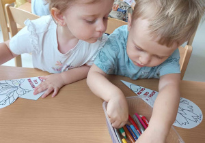 Nadia przygląda się jak Daniel wybiera z plastikowego prostokątnego pojemniczka jedną z kolorowych kredek.