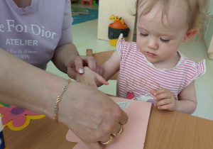 Opiekunka pomaga Mai przykleić na laurkę karteczkę z życzeniami dla Mamy.