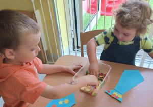 Maksymilian i Szymon wyciągają z plastikowego prostokątnego pojemniczka kolorowe aplikacje.