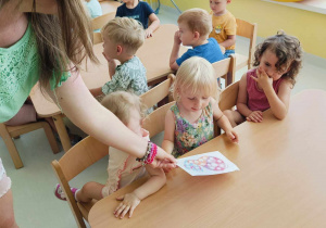 Barbara przygląda się, jak opiekunka daje Aleksandrze wyklejankę do wyklejenia.