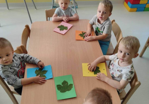 Fotografia dzieci siedzących przy stoliku przyklejających liście na kolorowe kartki.
