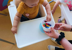 Opiekunka podaje dziewczynce styropianowy pojemniczek z bibułowymi kuleczkami.