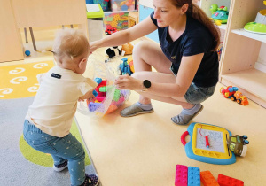 Dziewczynka razem z opiekunką wkładają do przezroczystego worka kolorowe klocki typu cegły.