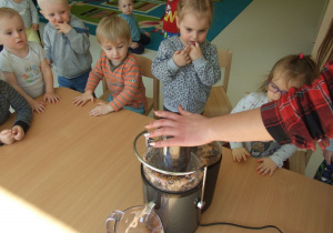Dzieci siedzące przy stole wraz z opiekunką wyciskają sok