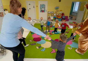 Miś Krzyś tańczy z dziećmi z grupy Żabki w kółeczku na dywanie.